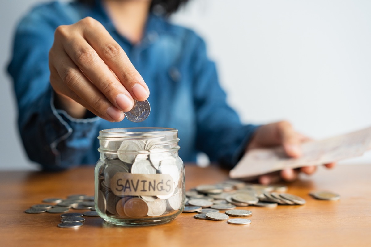 The woman hand is putting a coin in a glass bottle and a pile of coins on a brown wooden table,Investment business, retirement, finance and saving