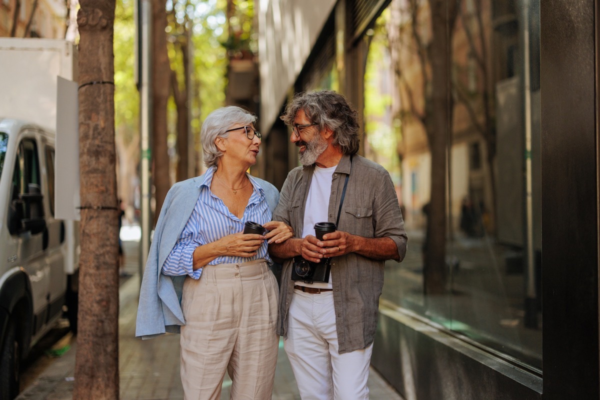 A cheerful senior couple is outside in the city walking on the street, socializing and drinking coffee.