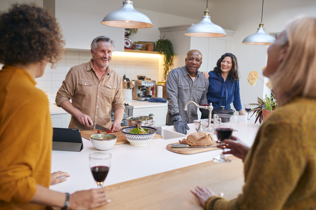 Group Of Mature Friends Meeting At Home Preparing Meal And Drinking Wine Together