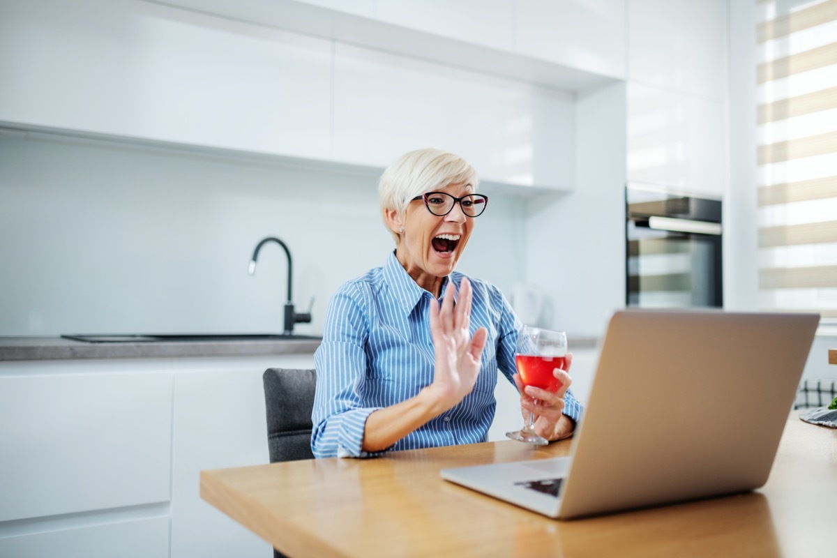 Happy smiling caucasian blonde senior woman sitting at dining table, drinking red wine, having video call over at laptop and waving.