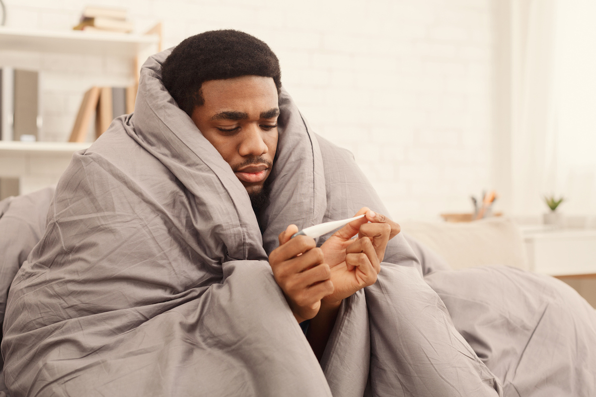 Ill man covered with blanket holding thermometer, having fever.