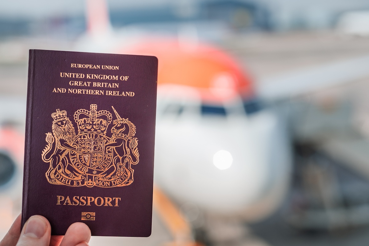 A biometric British passport being held up against a bright white and orange plane in the background