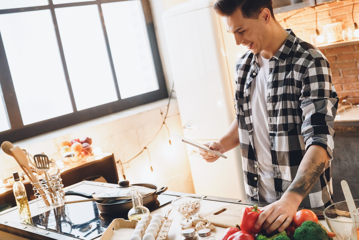 young adult man holding tablet smiling and cooking dinner