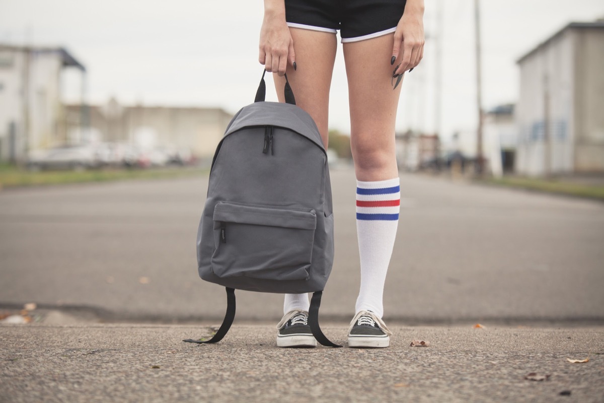 Girl holding generic backpack wearing black shorts and tube sock with tennis shoes standing on vacant street. - Image