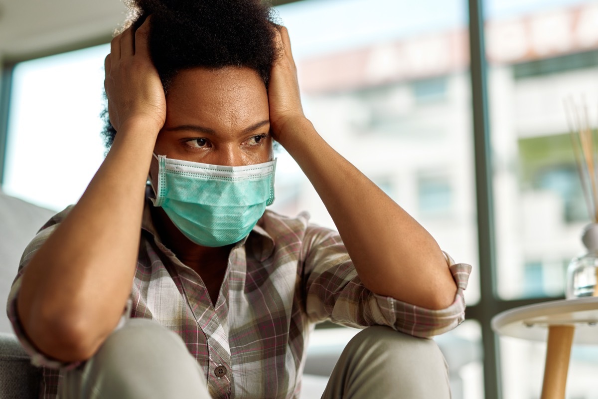 woman with face mask holding her head in pain and looking away at home