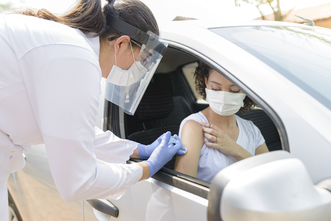 A young woman sitting in her car wearing a face mask receives a COVID-19 vaccination from a female healthcare worker wearing a face shield, face mask, and gloves.