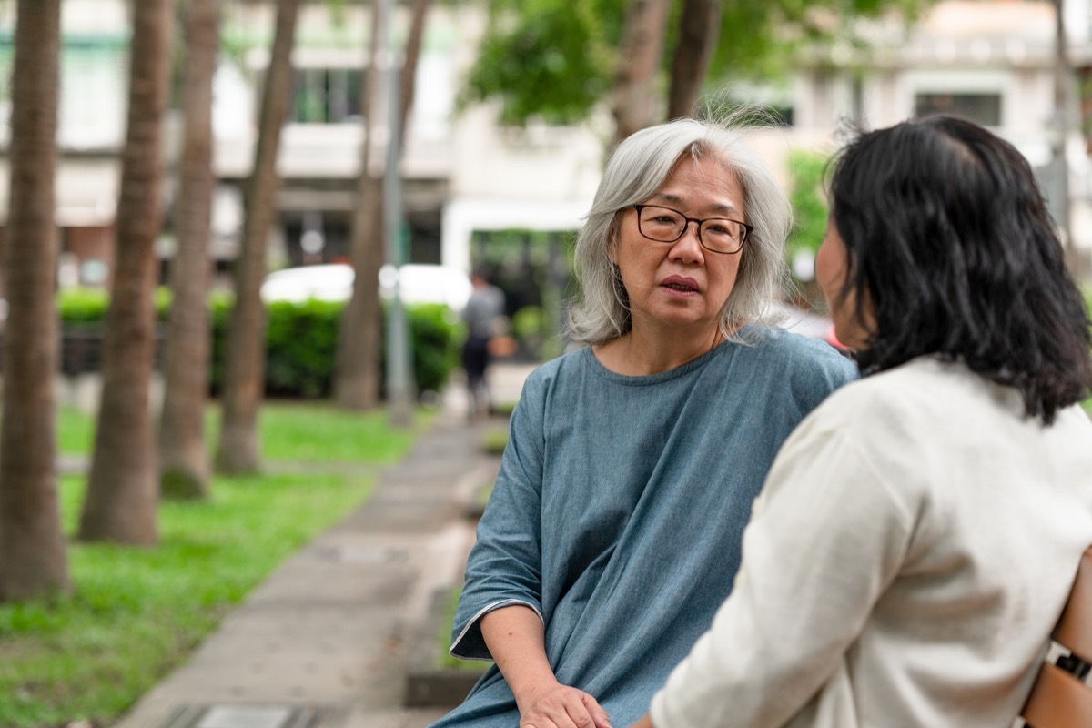 Two close senior friends are sitting on a park bench, spending time together talking about life and using social media.