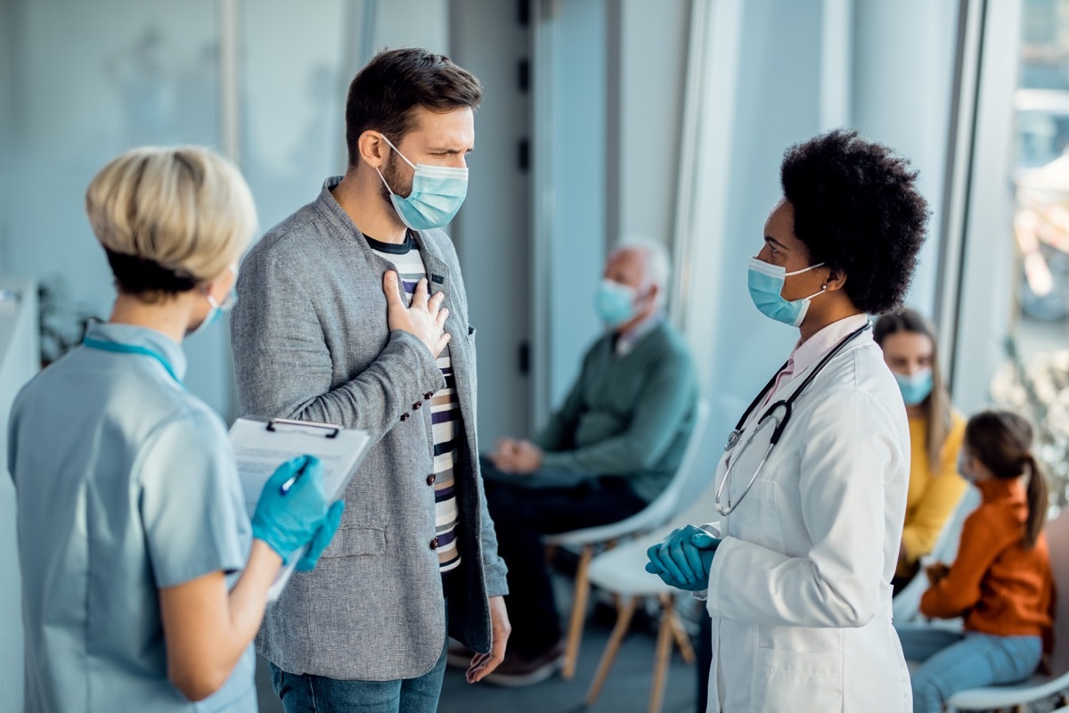 Distraught man complaining at chest pain while talking to doctor in a hallway at medical clinic during coronavirus pandemic.