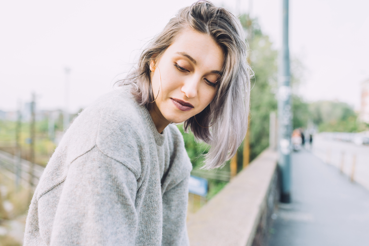 A young woman with purple-gray hair sits on a ledge and looks away.