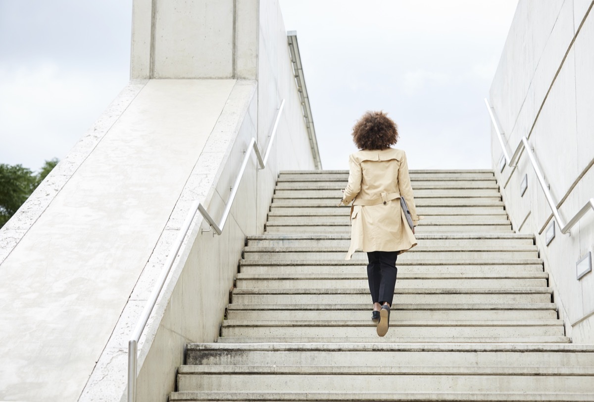 Full length rear view of businesswoman moving up steps. Low angle view of fashionable professional is holding folder. She is wearing long coat.