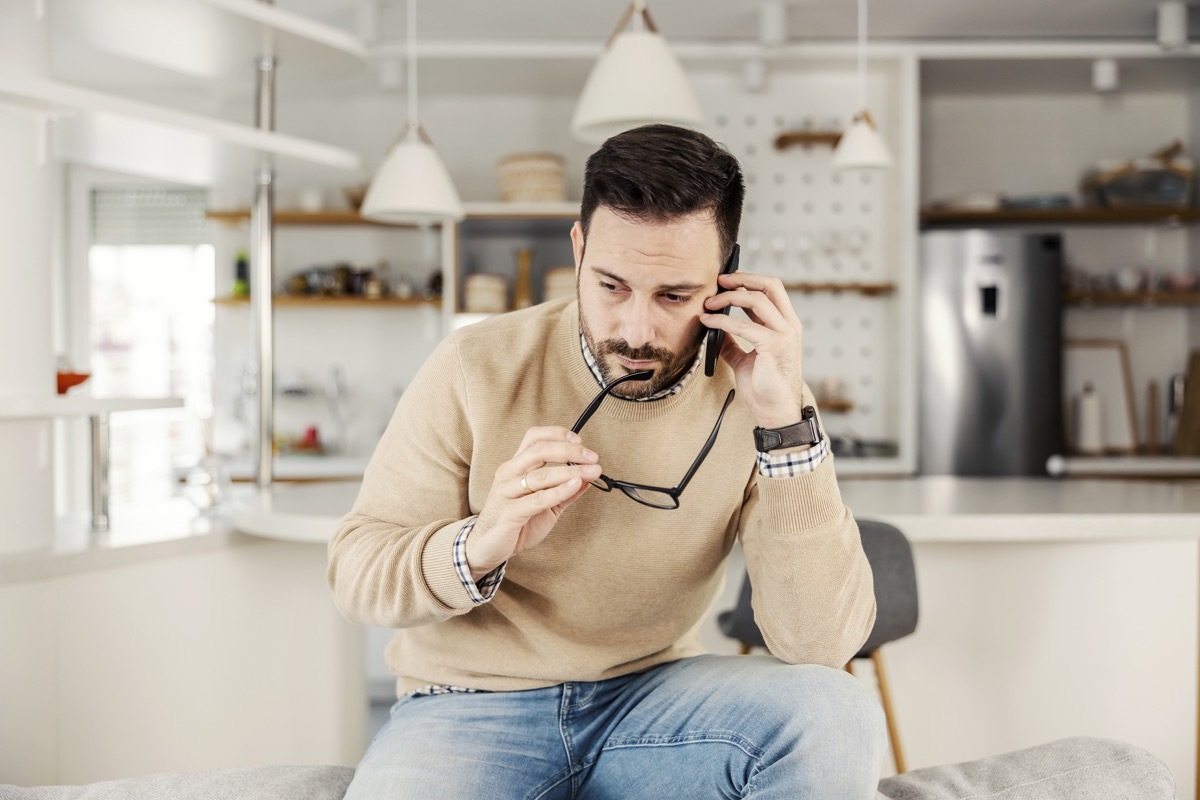 A worry man sitting at his cozy home, holding eyeglasses and talking on the phone.