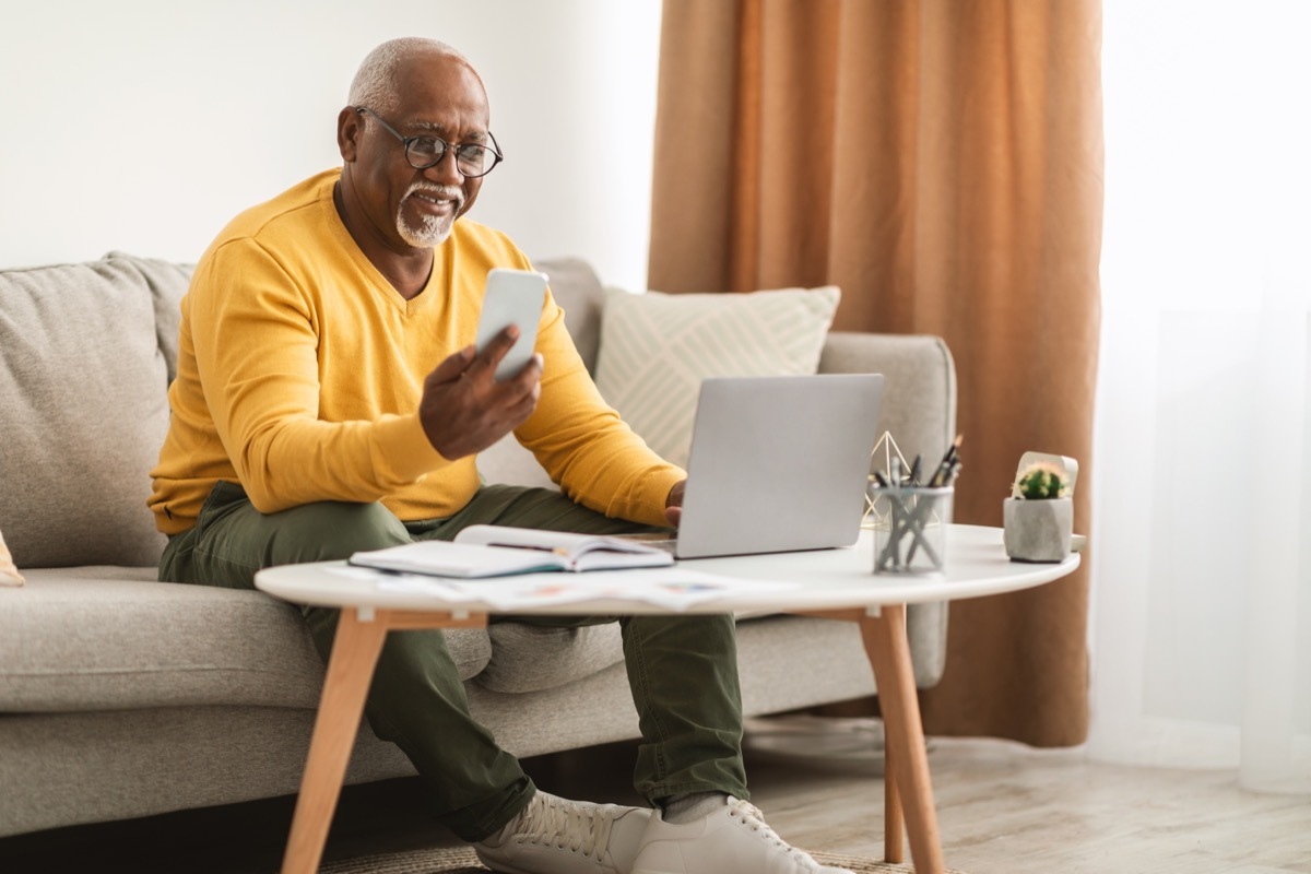 man smiling as he looks at his cell phone while sitting on the couch also using his laptop