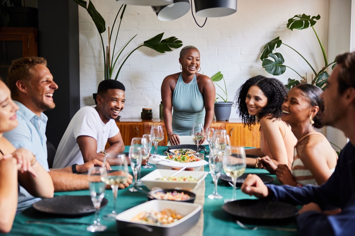 Young woman laughing at the head of a table while hosting a dinner party for a group of diverse young friends at her home