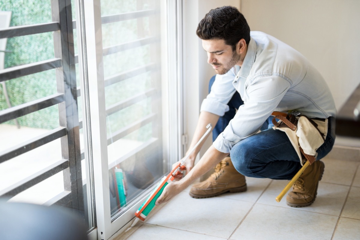 handyman wearing a tool belt and using a sealing gun to seal a glass door frame on a house