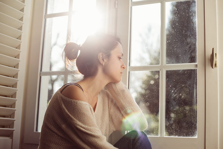 A Caucasian woman sits at a window with a strained look on her face