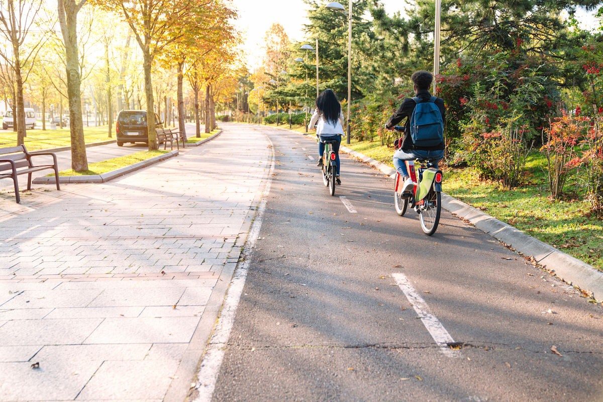 two teenagers young students man and woman riding along a bicycle path with a sharing electric bicycle e-bike in a beautiful park with many trees at sunset