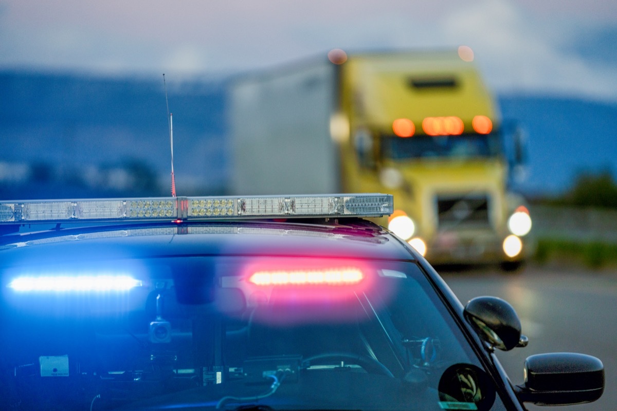 A patrol car with lights flashing is pulled off the shoulder to give a driver a ticket as a semi truck is driving by