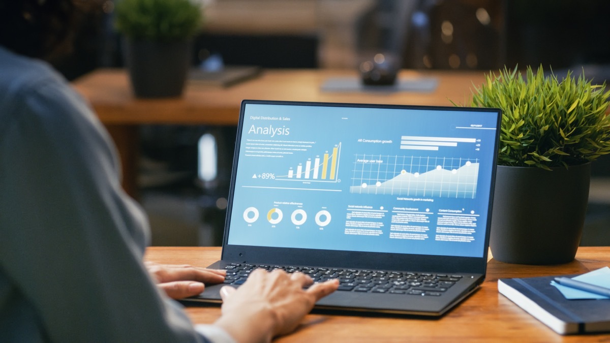 Female Analyst at Her Desk Works on a Laptop Showing Statistics, Graphs and Charts. She Works on the Wooden Table in Creative Office. Over the Shoulder Footage.