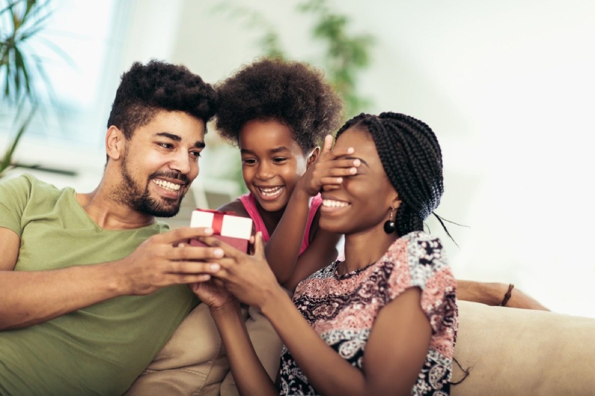 black mother, father, and daughter opening present on couch