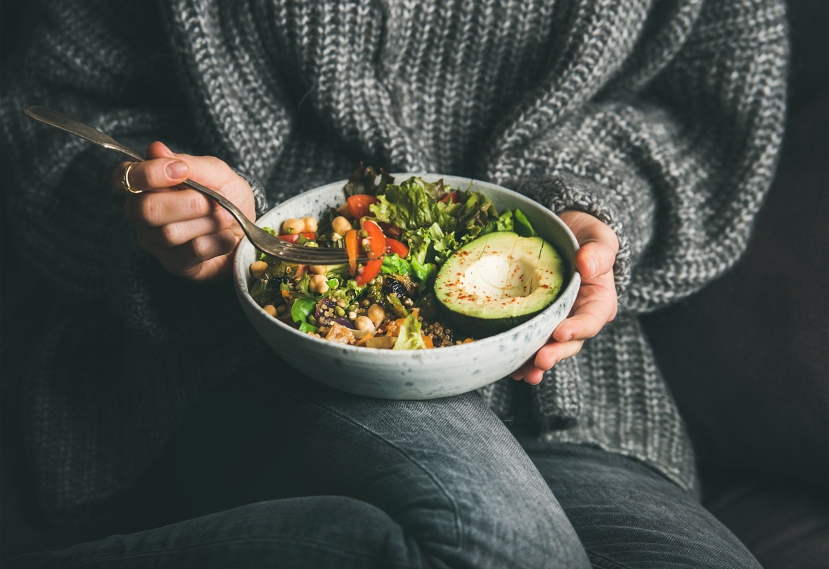 Healthy vegetarian dinner. Woman in grey jeans and sweater eating fresh salad, avocado half, grains, beans, roasted vegetables from Buddha bowl