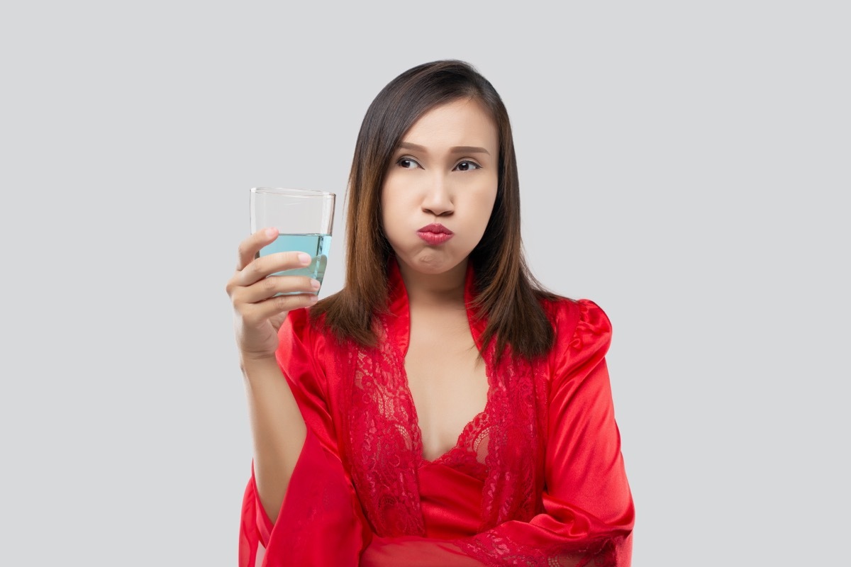 Woman in silk nightwear wearing red robe rinsing mouth with mouthwash