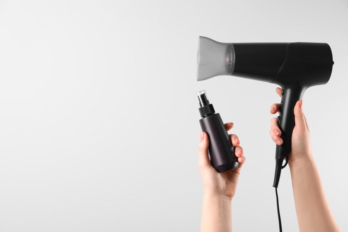 Woman holding spray bottle with thermal protection and hairdryer on white background, closeup. Space for text