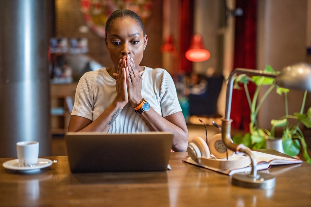 woman holds her hand to her face in shock, reading laptop inside