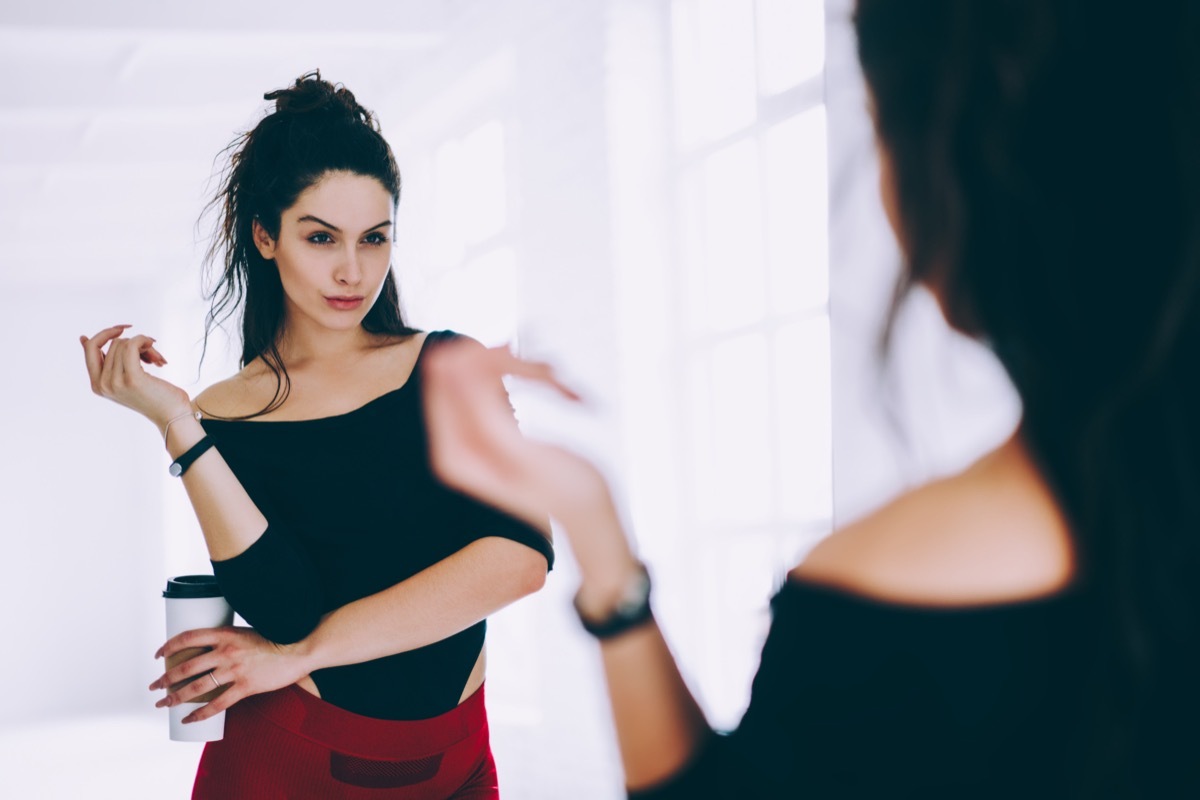 A woman looks at herself in the mirror wearing a black tank top.