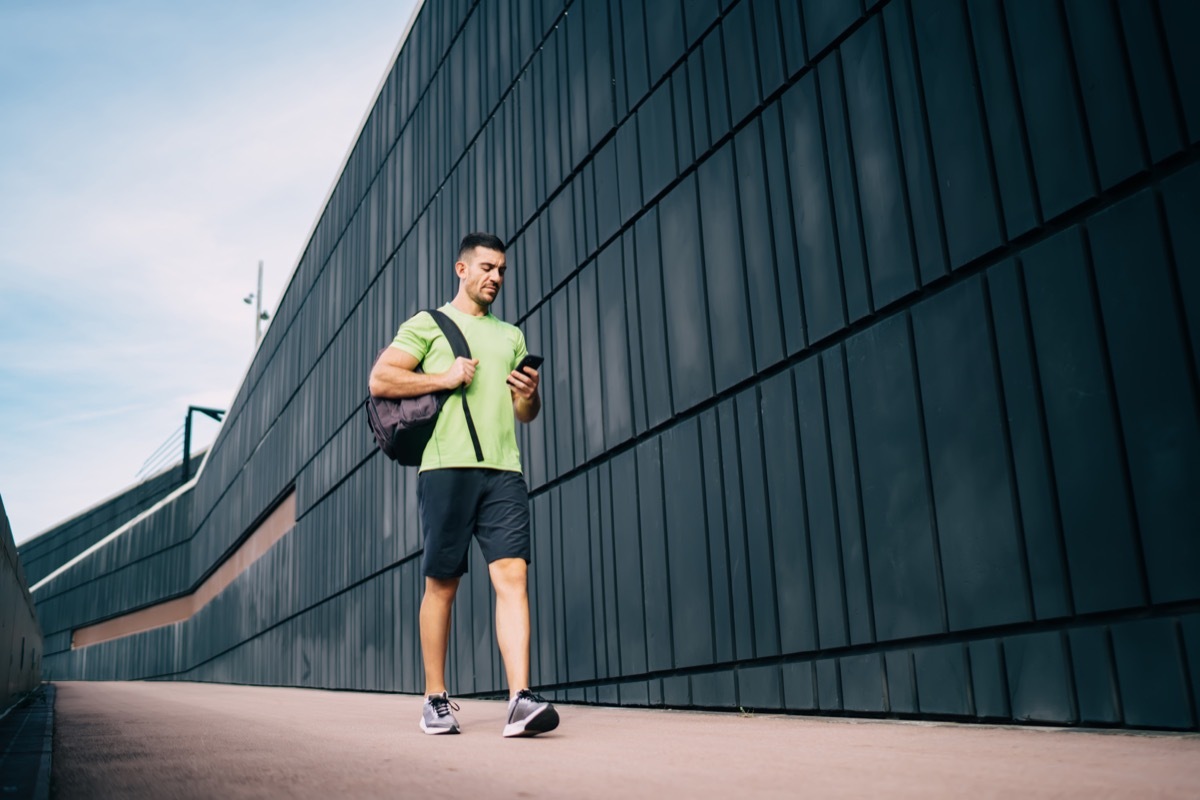 Male in sportswear with backpack walking down road past wall of sports building while using mobile phone