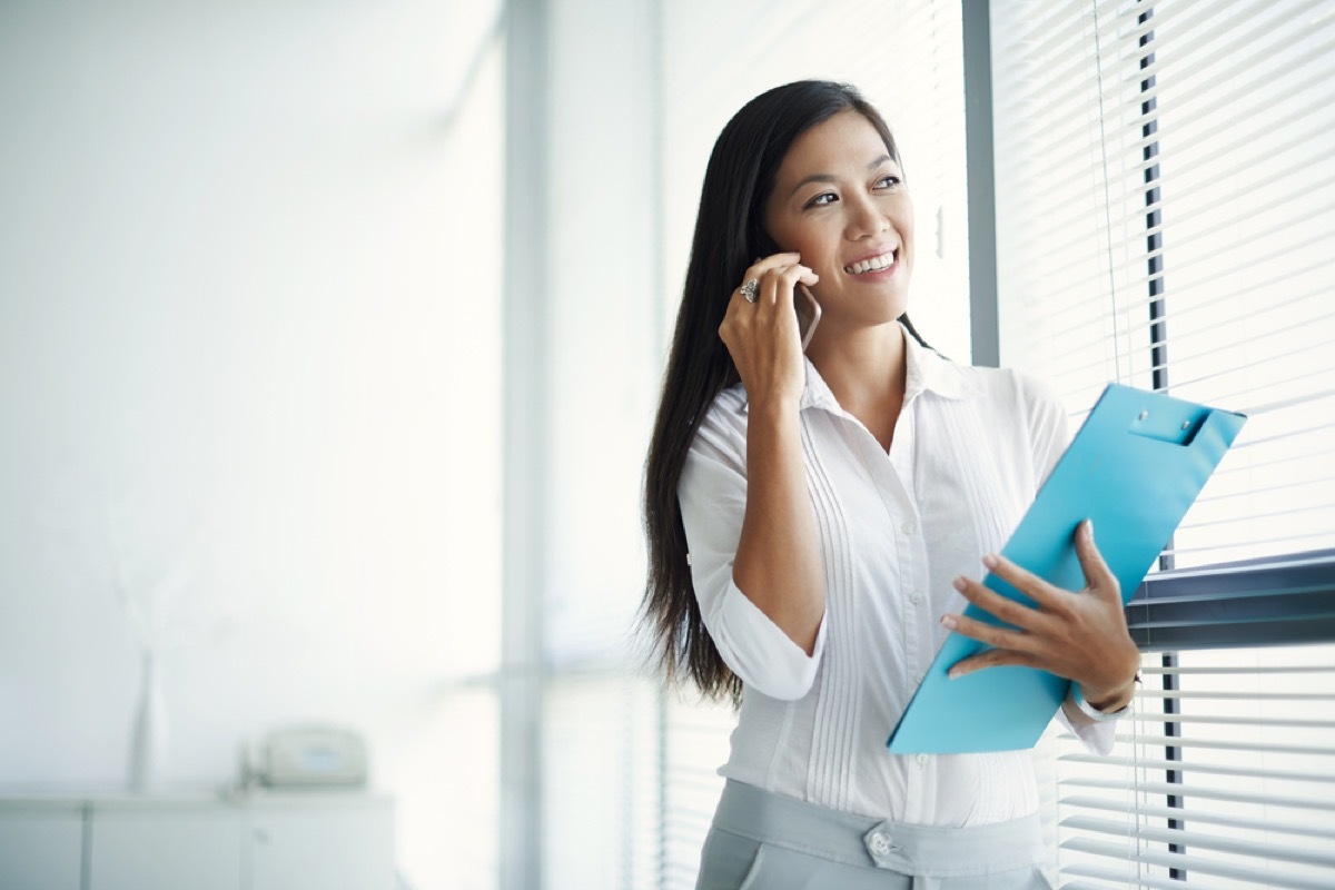 young asian woman carrying clipboard and talking on smartphone, reach a customer service rep