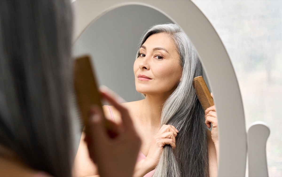 Senior attractive middle 50 years aged asian woman with gray hair looking at mirror reflection combing tangled gray hair. Alopecia hair loss prevention treatment after menopause advertising concept.