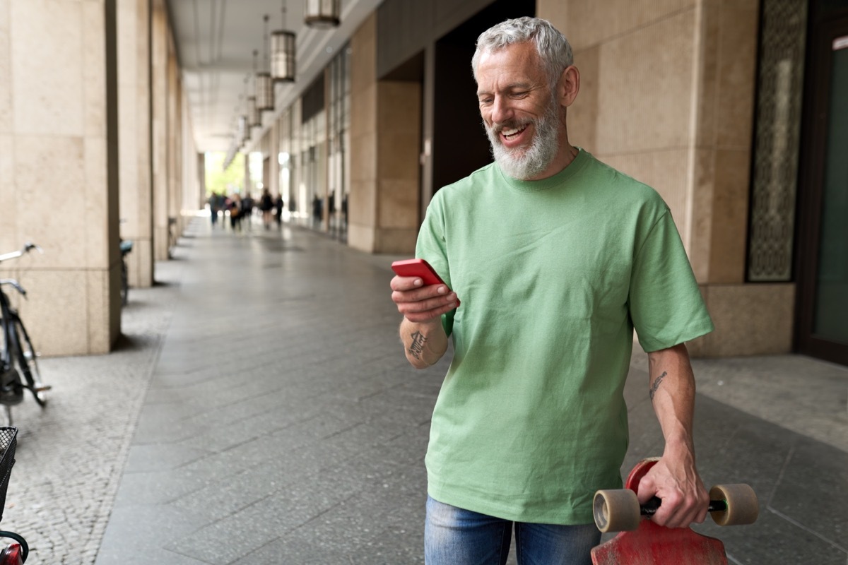 Man carrying a skateboard and looking down at his phone laughing