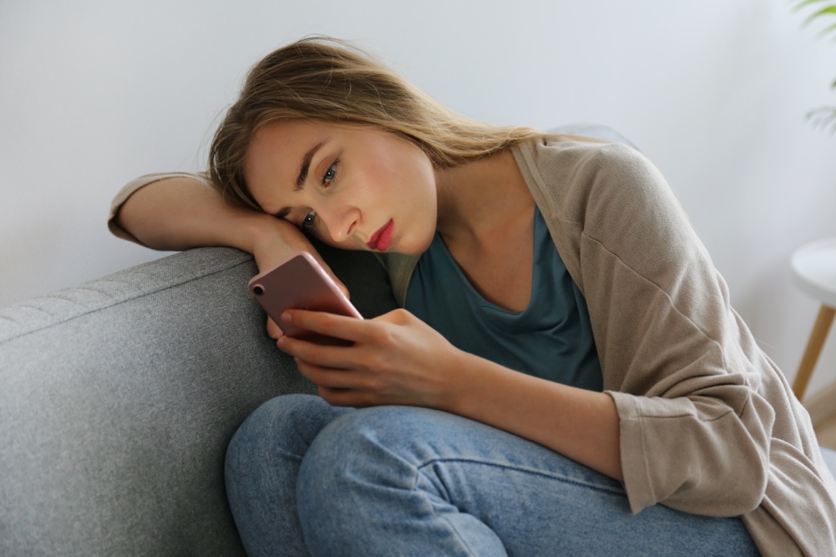 woman with depressed facial expression sitting on grey textile couch holding her phone