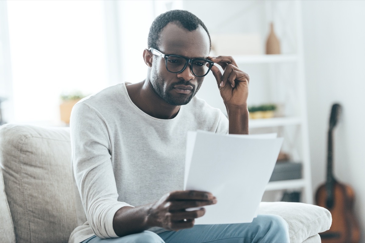 man reading documents and touching eyeglasses with his hand while sitting on the sofa at home