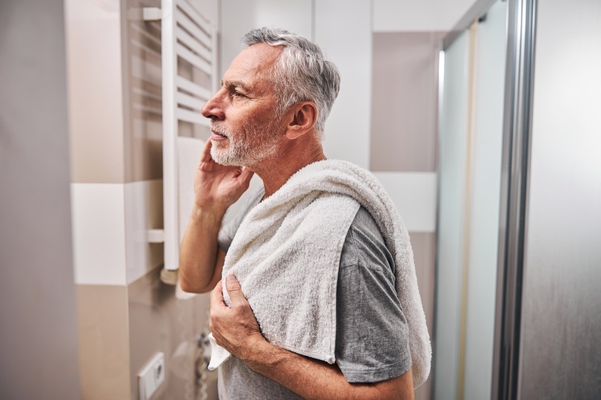 Side-view photo of a serious aged man looking in the mirror while standing in the bathroom with a towel