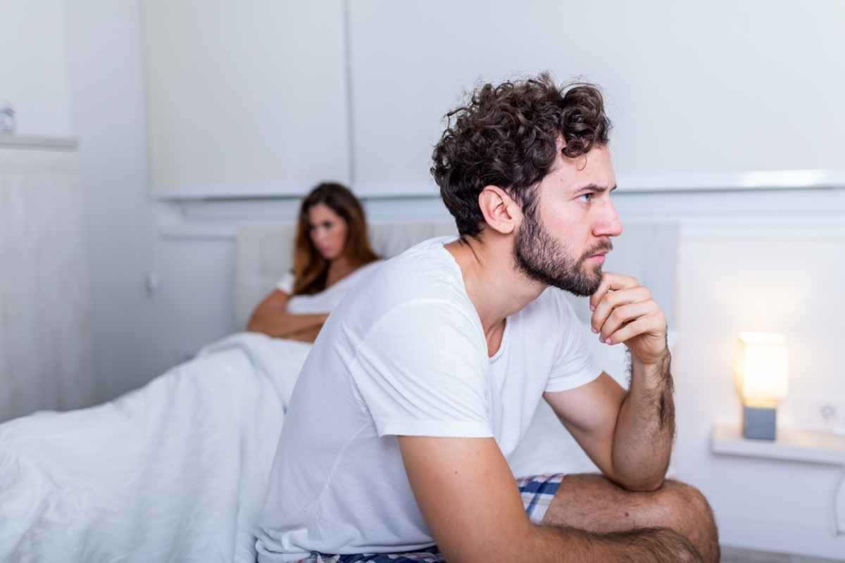 Sad man sitting on a bed, girlfriend in the background.