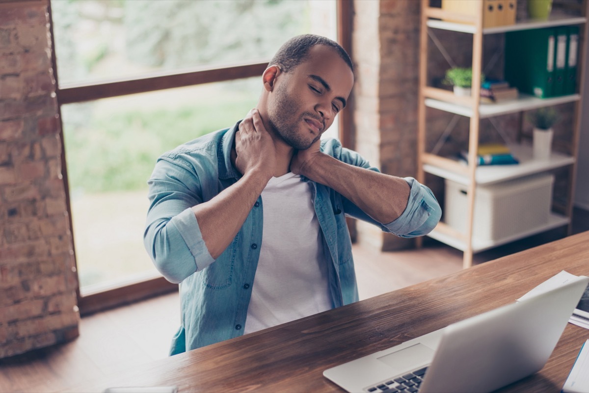 Young frustrated afro american freelancer is having a strong pain in neck. He is at home office, sitting at his modern work station, suffering, massaging it