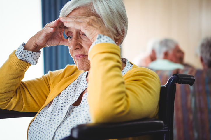 A senior woman sitting in a wheelchair holding her head looking confused and worried