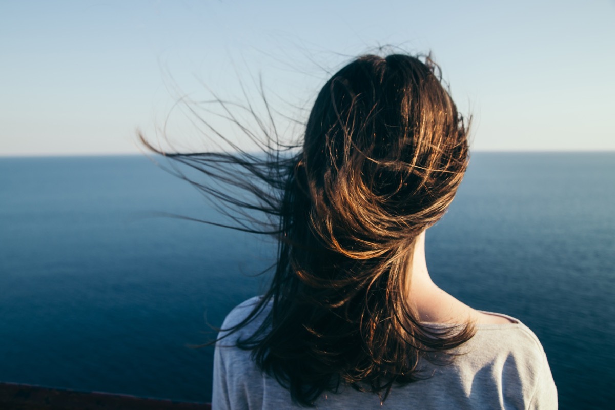 Woman with dark hair stands on a top cliff over blue sea view while wind.