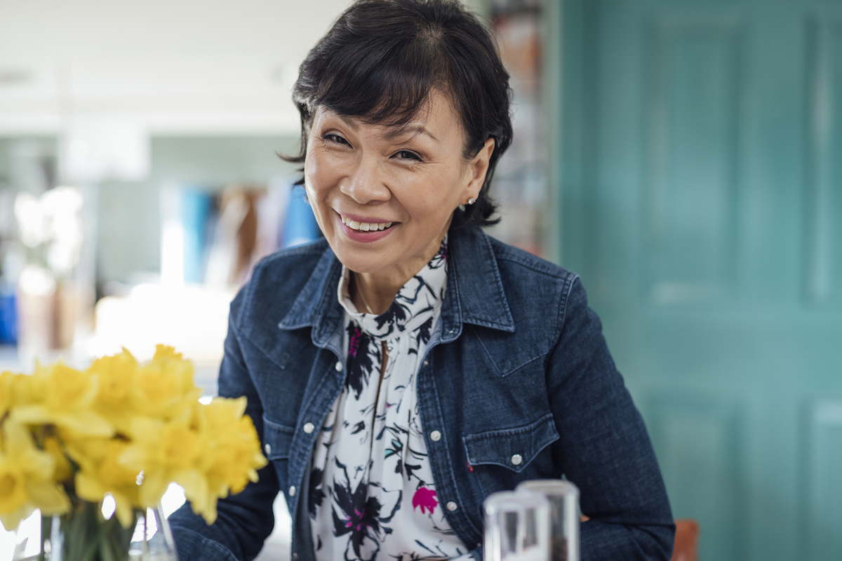 Mature woman smiling wearing a jean jacket and a blouse next to a vase of daffodils