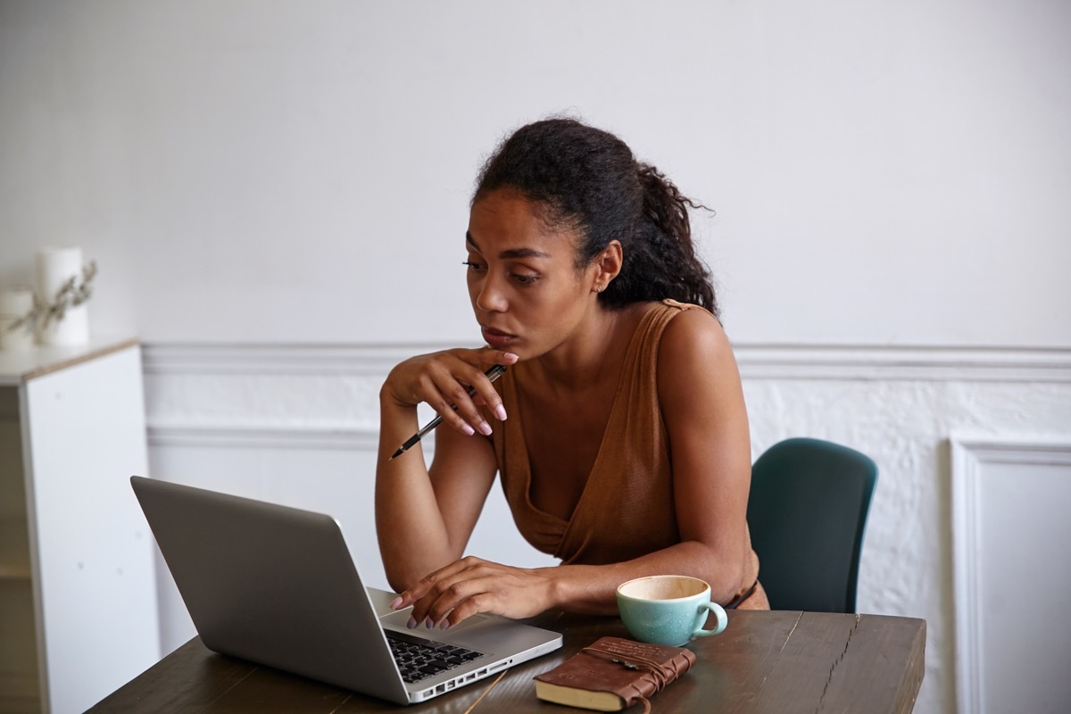 Beautiful dark skinned businesswoman with casual hairstyle working on her laptop, looking at screen with concentrated face and touching chin with hand