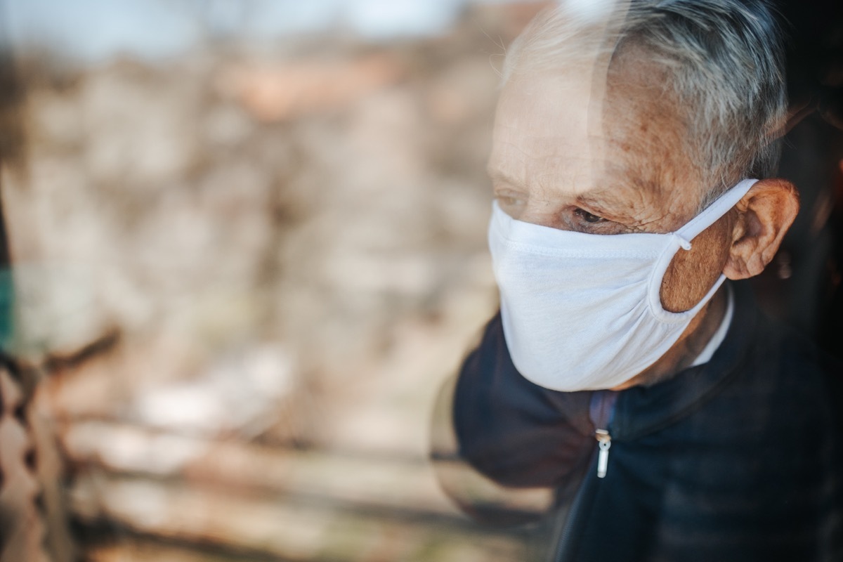 Senior man in home quarantine wearing a mask and looking through the window, during coronavirus COVID-19 epidemic.