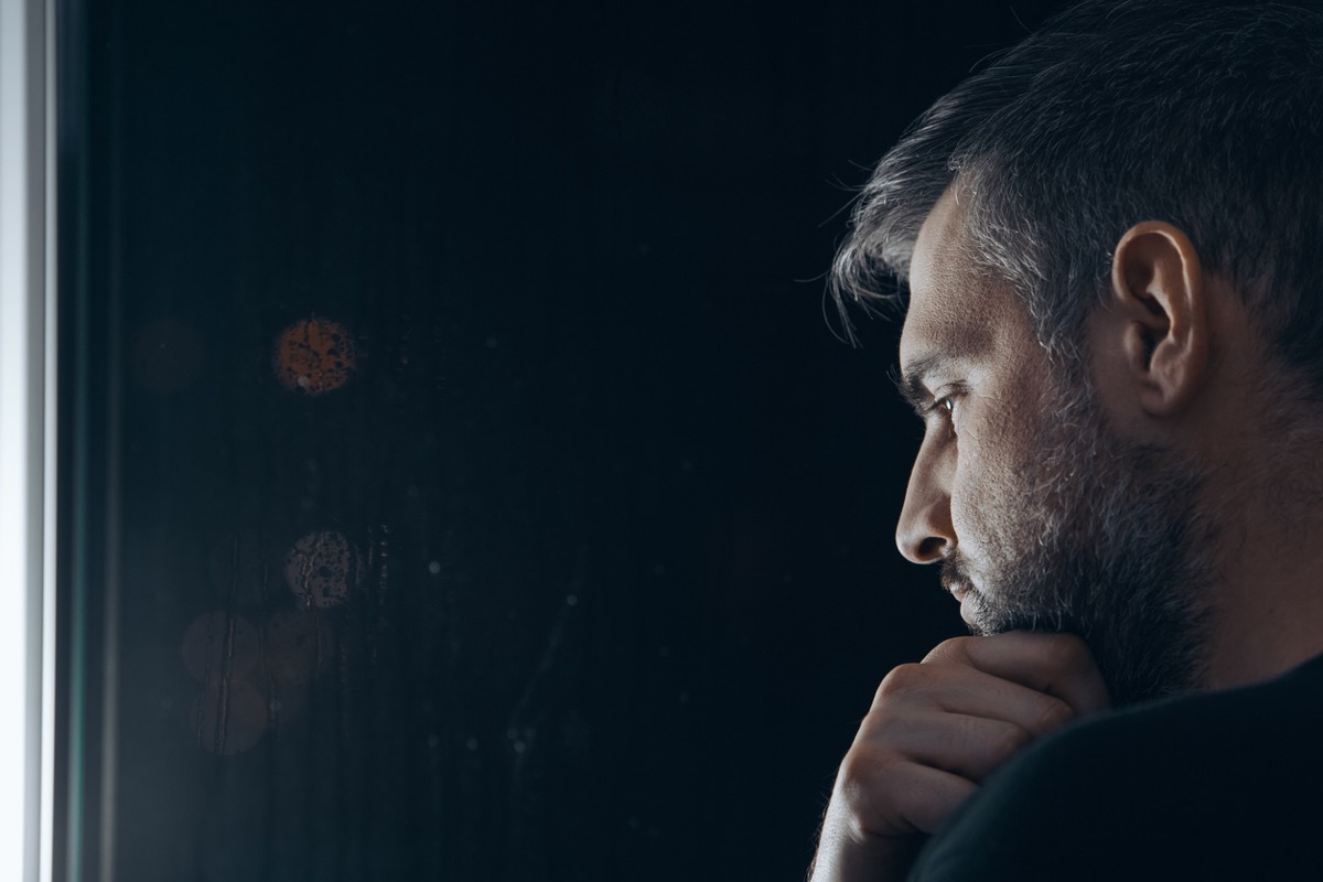 Man with beard holding his chin, standing beside a window at night