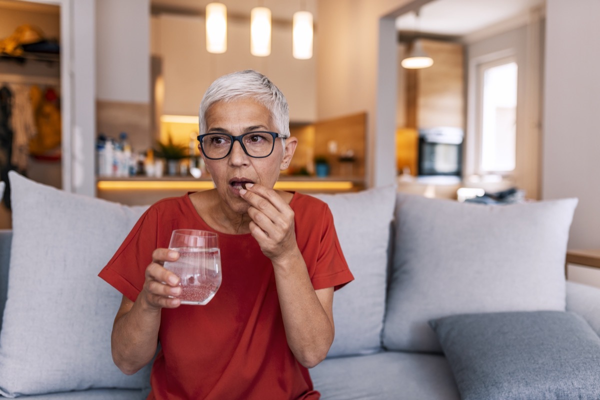 Mature woman's morning routine - holding medicine and water. Photo of Mature woman with pill and glass of water at home. Mature woman sitting on bed, suffering from depression. Woman takes pill with omega-3 and holding a glass of fresh water.