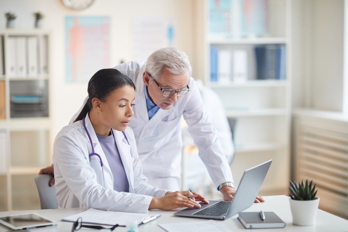 Senior doctor standing and looking at laptop computer while young nurse sitting at the table and typing on laptop at office