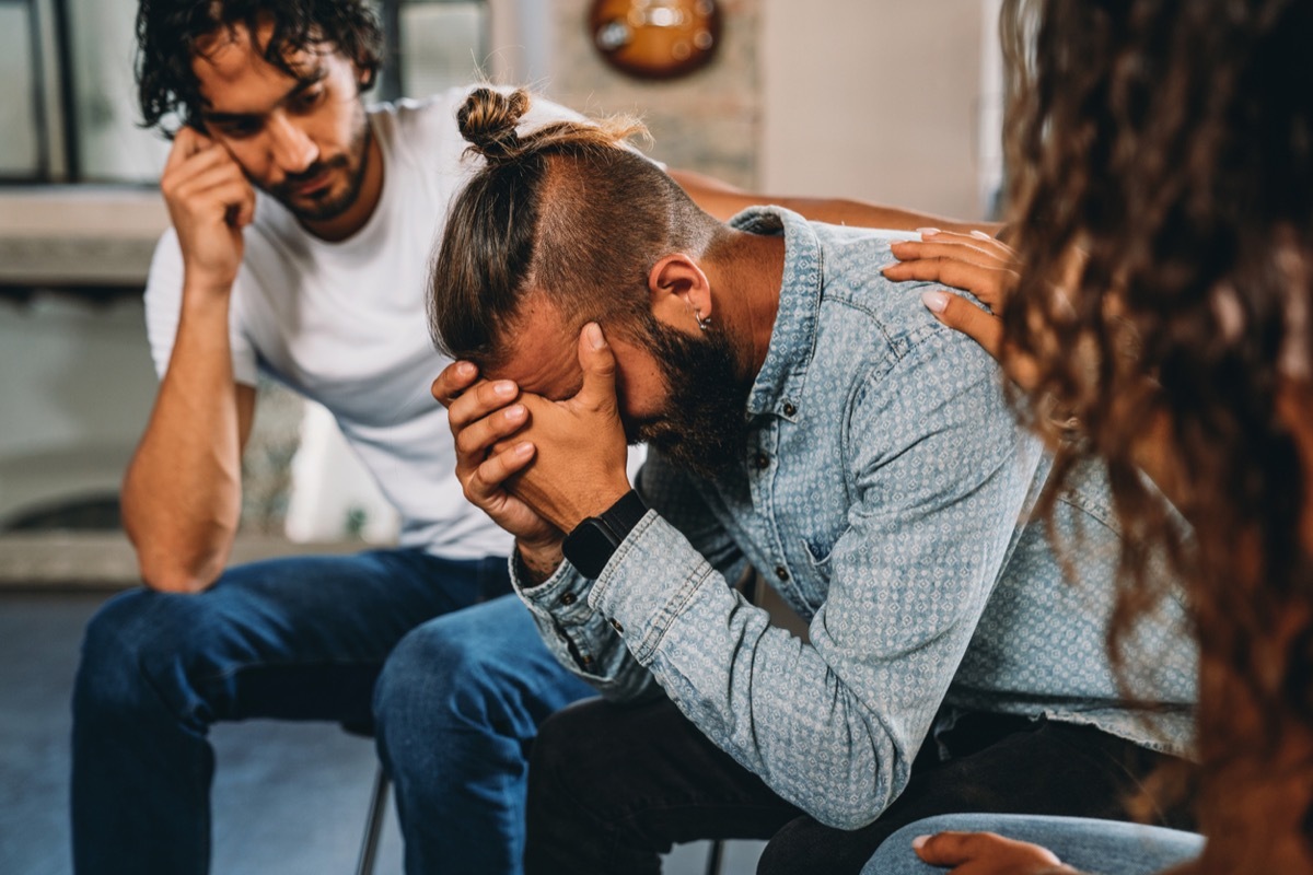 People are comforting a man during a group therapy session. Mental health concept in a loft with big windows. Healthcare and medicine concept. Group of people sitting in a circle are participating in a support meeting, talking with each other.