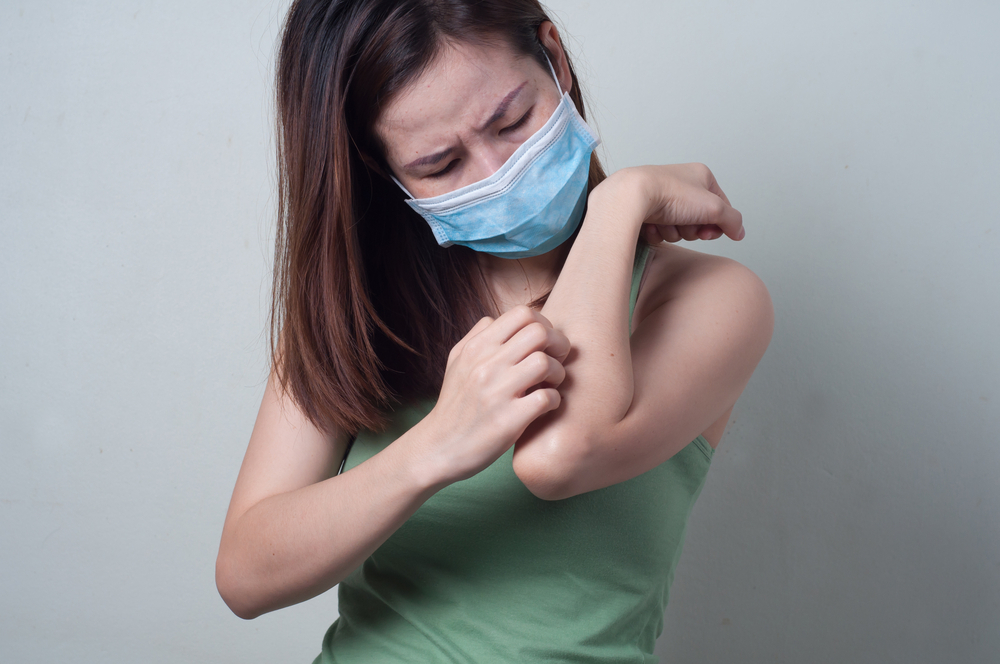 young woman scratching arm while wearing surgical mask